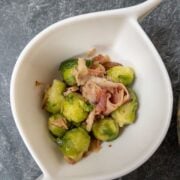 Brussels sprouts with fried bacon and chestnuts in a curved white bowl on a marble surface.