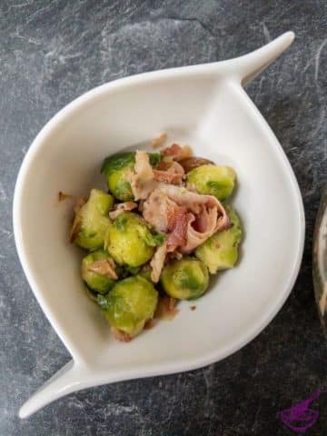 Brussels sprouts with fried bacon and chestnuts in a curved white bowl on a marble surface.