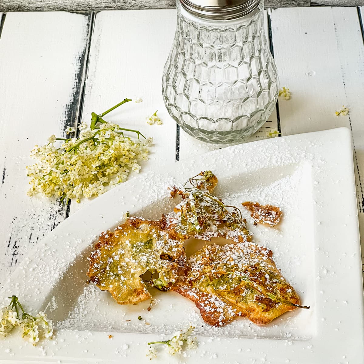 Baked elderflowers on white plate. Muffineer in the back and one fresh elderflower bunch beside.