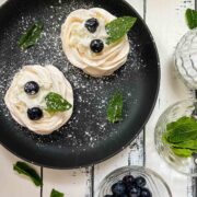 Meringue nests filled with whipped cream, decorated with blueberries and mint leaves, and dusted with powdered sugar. Placed on black serving plate.