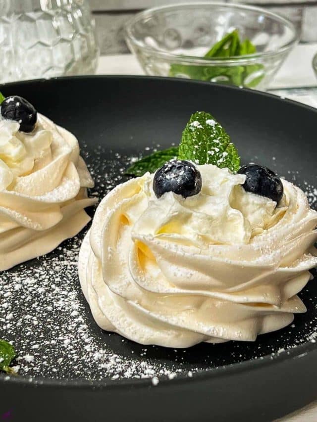 Meringue nests filled with whipped cream, decorated with blueberries and mint leaves, and dusted with powdered sugar. Placed on black serving plate.