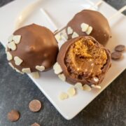 Three lotus biscoff cake pops with bite marks on a white plate. Surrounded by milk chocolate chips and white chocolate hearts.