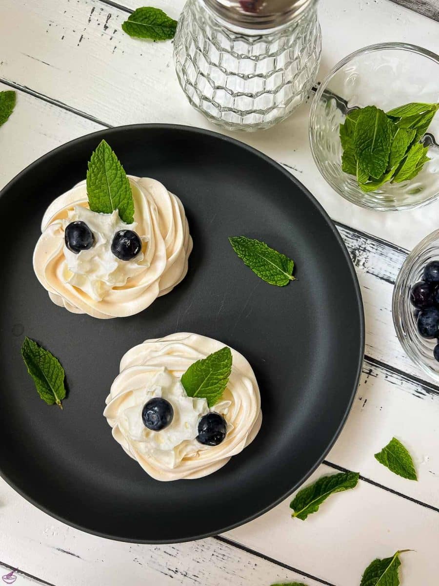 Meringue nests on a black serving plate, filled with whipped cream, and garnished with blueberries and mint leaves. 