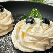 Meringue nests filled with whipped cream, decorated with blueberries and mint leaves, and dusted with powdered sugar. Placed on black serving plate.