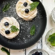 Meringue nests filled with whipped cream, decorated with blueberries and mint leaves, and dusted with powdered sugar. Placed on black serving plate.