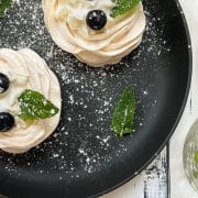 Meringue nests filled with whipped cream, decorated with blueberries and mint leaves, and dusted with powdered sugar. Placed on black serving plate.
