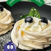 Meringue nests filled with whipped cream, decorated with blueberries and mint leaves, and dusted with powdered sugar. Placed on black serving plate.