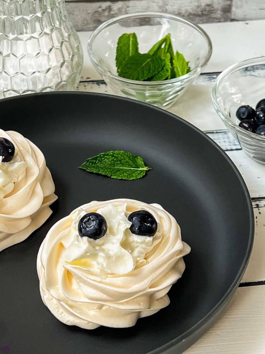 Meringue nests on a black serving plate, filled with whipped cream, and garnished with blueberries.