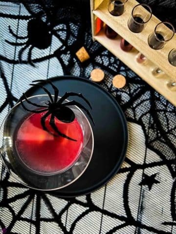 Blood red Halloween cocktail in glass decorated with spider. Black serving plate and spider web decoration.