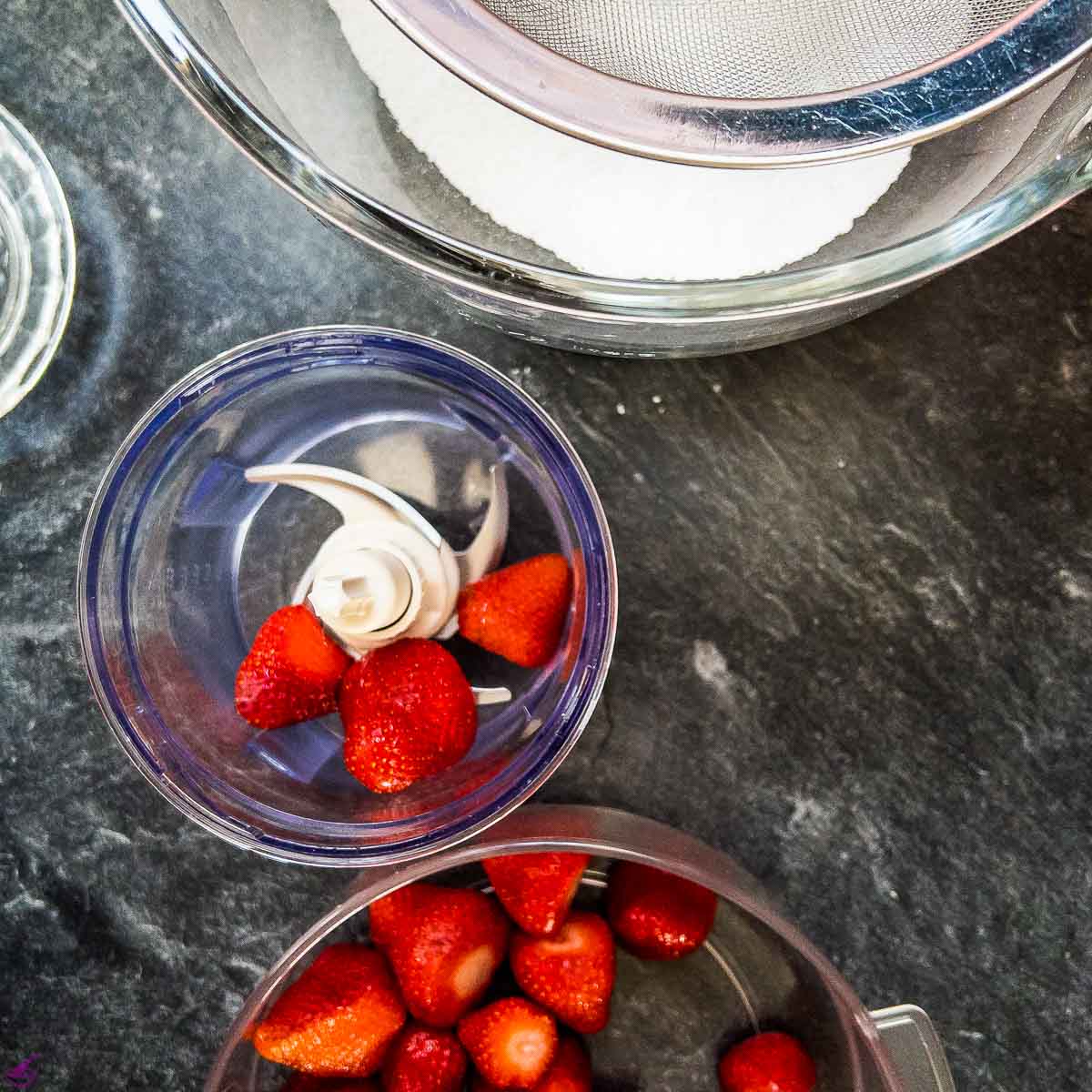 Strawberries placed in electric chopper next to a plastic mixing bowl filled with strawberries.