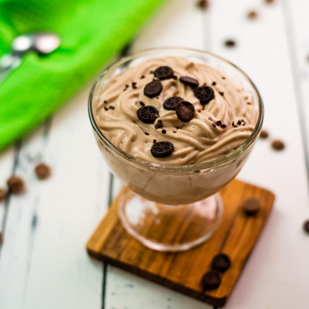 Beautifully textured chocolate cornstarch pudding in a dessert glass. Placed on a wooden coaster on a white wooden floor. Decorated with small pieces of cookies.