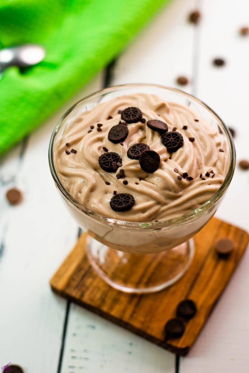 Beautifully textured chocolate cornstarch pudding in a dessert glass. Placed on a wooden coaster on a white wooden floor. Decorated with small pieces of biscuit.