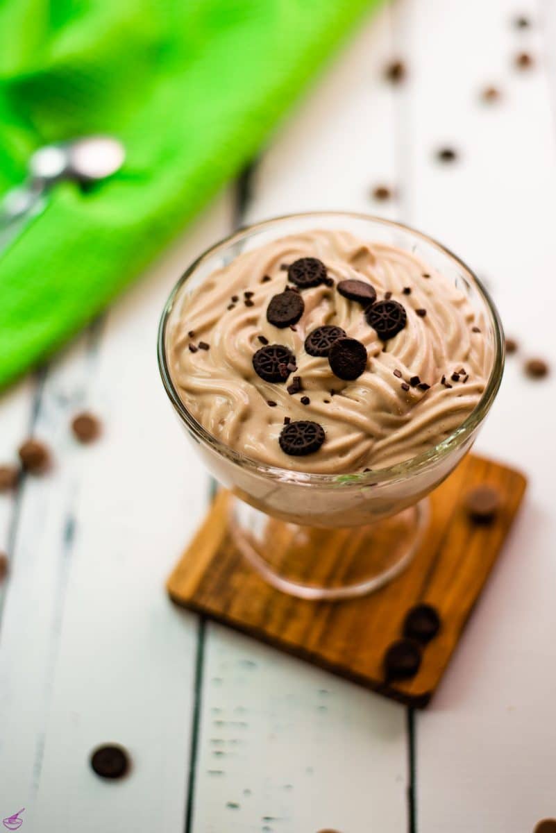 Beautifully textured chocolate cornstarch pudding in a dessert glass. Placed on a wooden coaster on a white wooden floor. Decorated with small pieces of biscuit.