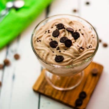 Beautifully textured chocolate cornstarch pudding in a dessert glass. Placed on a wooden coaster on a white wooden floor. Decorated with small pieces of biscuit.