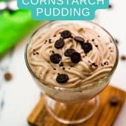 Beautifully textured chocolate cornstarch pudding in a dessert glass. Placed on a wooden coaster on a white wooden floor. Decorated with small pieces of cookies.