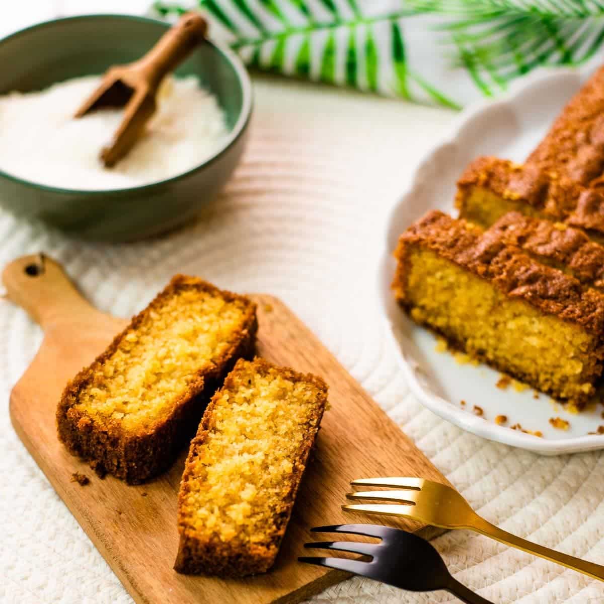 Two slices of coconut pound cake on a beautiful wooden board. In the background, you will find coconut flakes in a bowl and a sliced coconut pound cake.