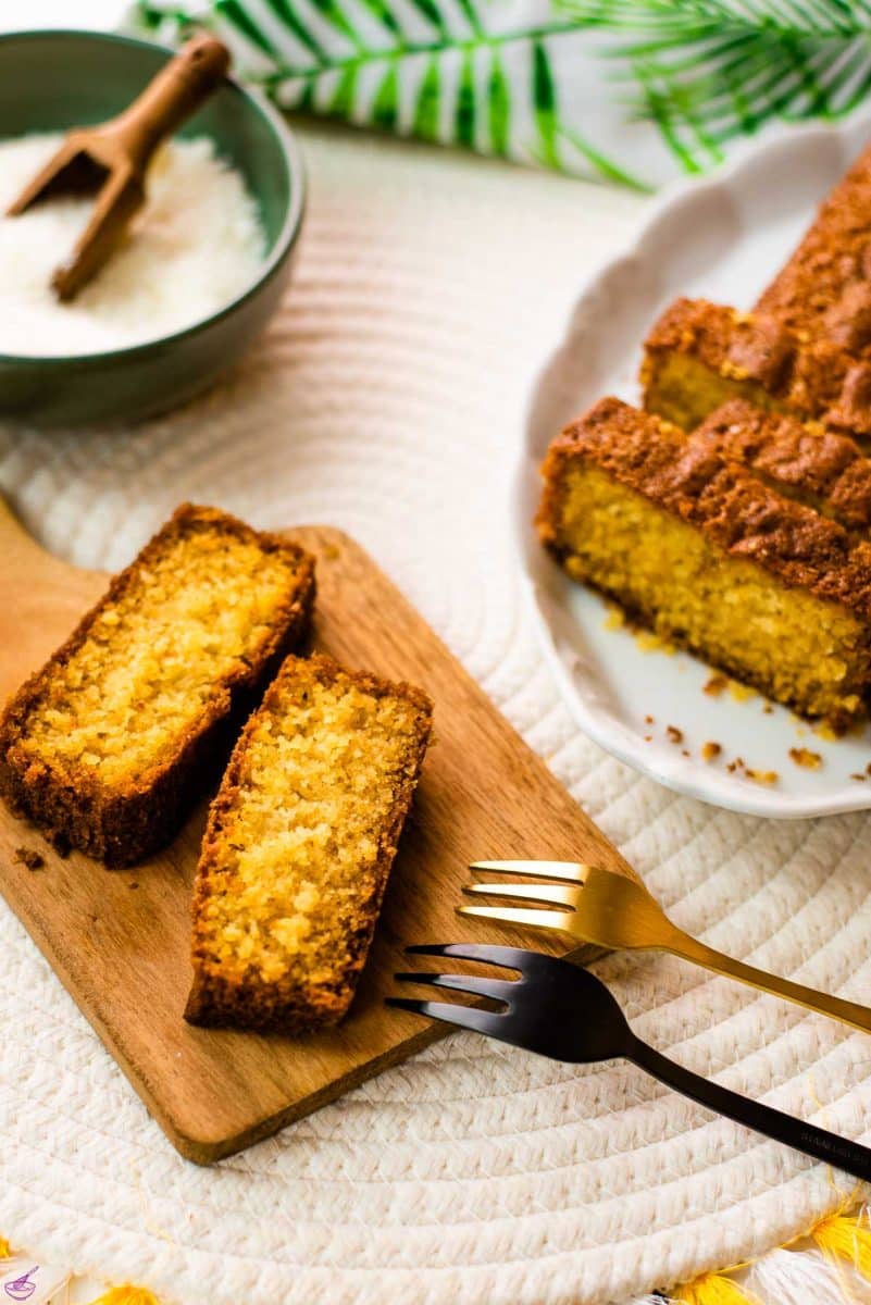 Two slices of coconut pound cake on a beautiful wooden board. In the background, you will find coconut flakes in a bowl and a sliced coconut pound cake.