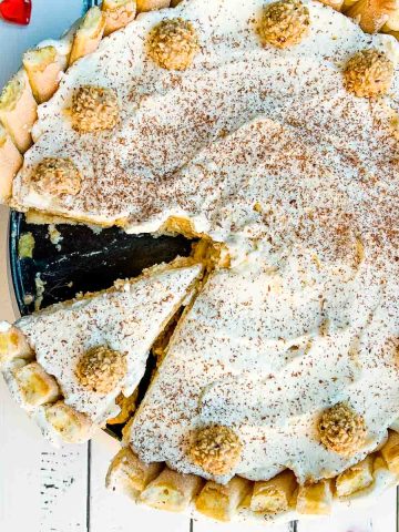 Delicious almond cream cake garnished with whipped cream, grated chocolate, and Giotto. It stands on a white wooden background surrounded by red glass hearts.