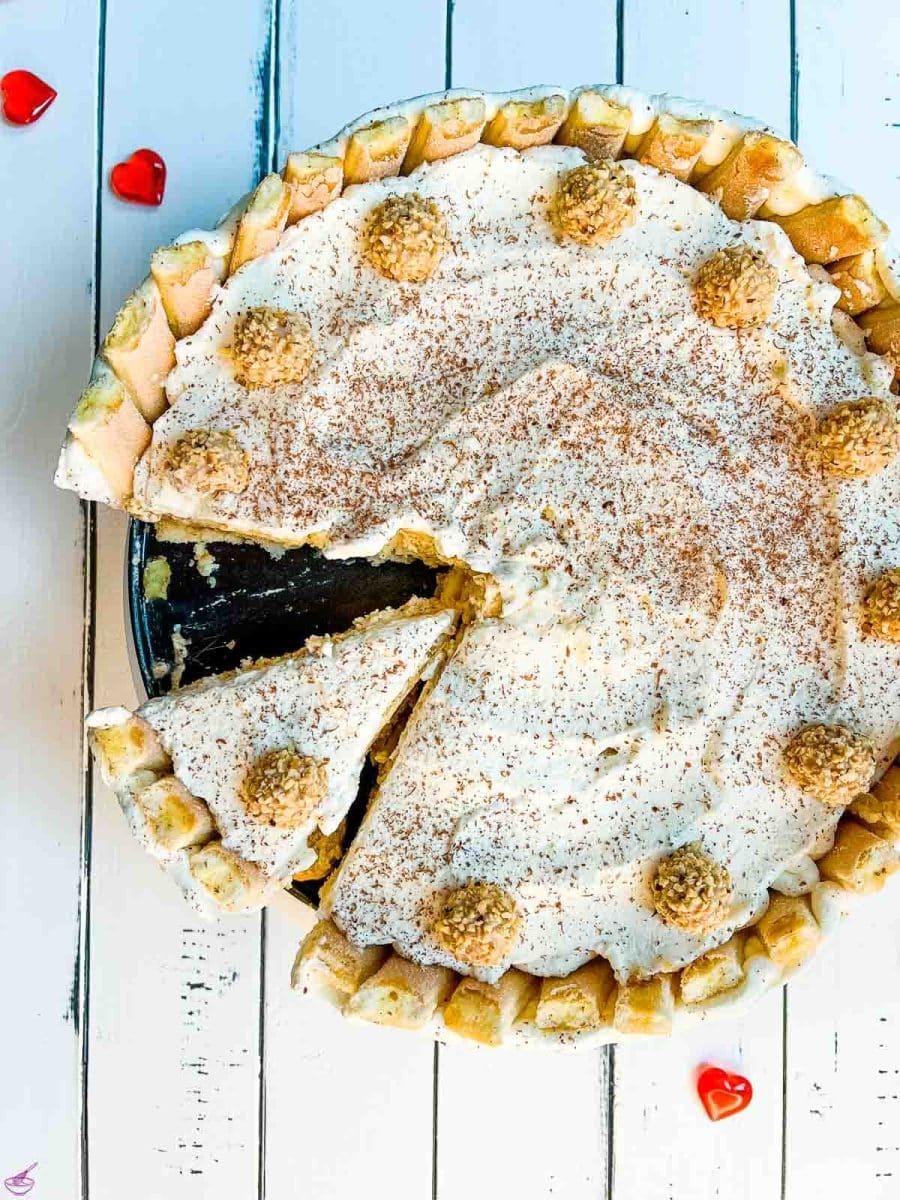 Delicious almond cream cake garnished with whipped cream, grated chocolate, and Giotto. It stands on a white wooden background surrounded by red glass hearts.