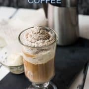 Beautiful glass filled with delicious vanilla iced coffee. Topped with chocolate-flavored whipped cream. It stands on a black slate against a light background.