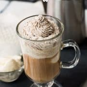 Beautiful glass filled with delicious vanilla iced coffee. Topped with chocolate-flavored whipped cream. It stands on a black slate against a light background.