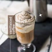 Beautiful glass filled with delicious vanilla iced coffee. Topped with chocolate-flavored whipped cream. It stands on a black slate against a light background.