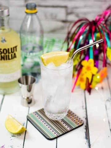 Beautiful glass filled with vanilla vodka tonic, on colorful coaster placed on white wooden board.