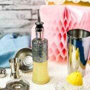 Bottle filled with freshly squeezed lemon juice fitted with a pouring spout, surrounded by cocktail making tools.