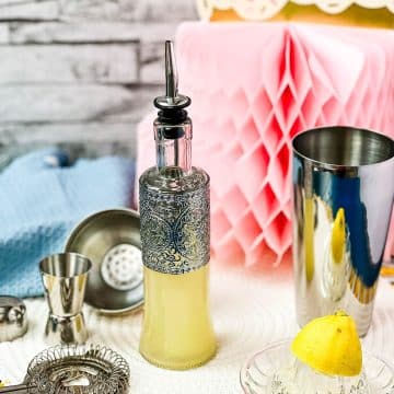 Bottle filled with freshly squeezed lemon juice fitted with a pouring spout, surrounded by cocktail making tools.