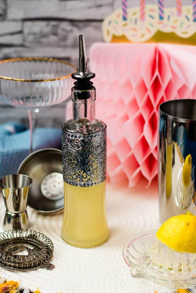 Bottle filled with freshly squeezed lemon juice fitted with a pouring spout, surrounded by cocktail making tools.