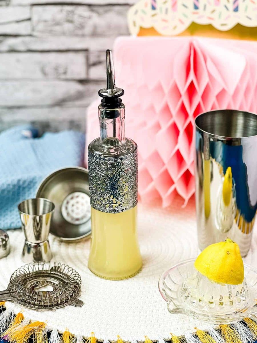 Bottle filled with freshly squeezed lemon juice fitted with a pouring spout, surrounded by cocktail making tools.