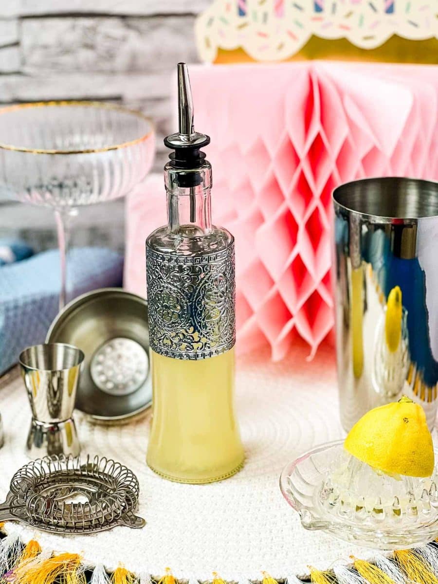 Bottle filled with freshly squeezed lemon juice fitted with a pouring spout, surrounded by cocktail making tools.