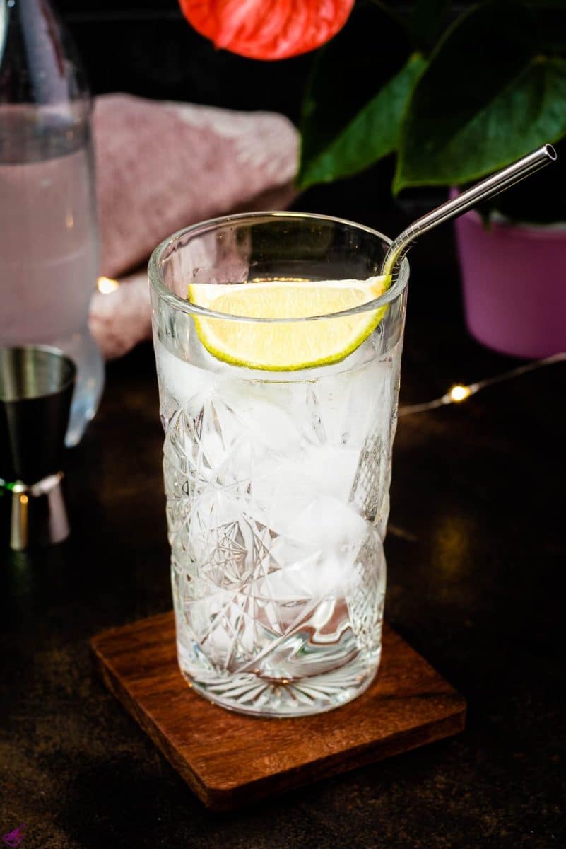 Highball glass filled with gin and tonic, garnished with lime, placed on wooden coaster.