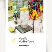 Beautiful glass filled with vanilla vodka tonic, on colorful coaster placed on white wooden board.