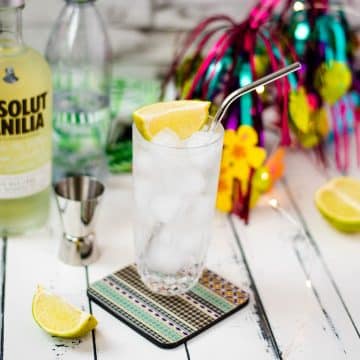 Beautiful glass filled with vanilla vodka tonic, on colorful coaster placed on white wooden board.