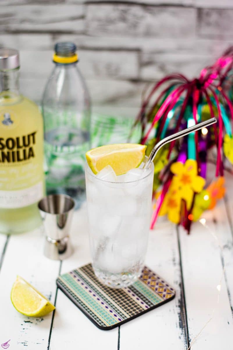 Beautiful glass filled with vanilla vodka tonic, on colorful coaster placed on white wooden board.