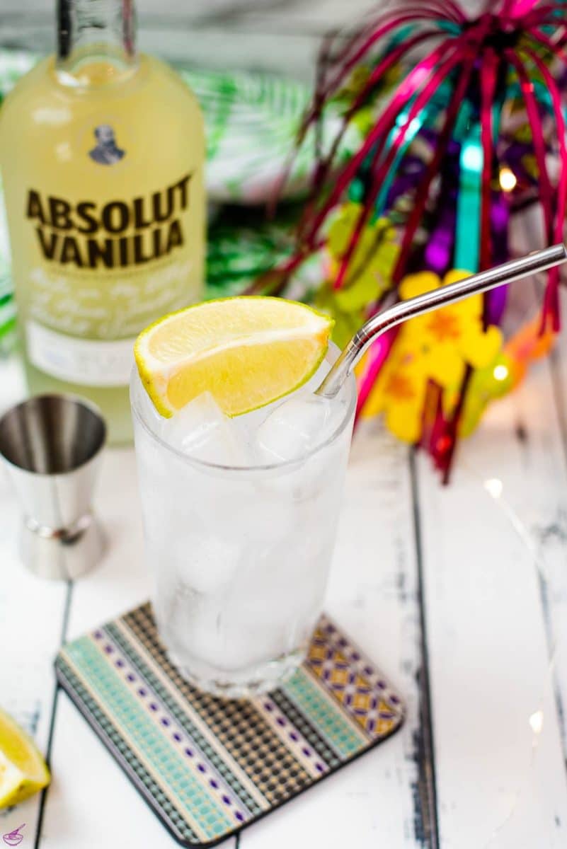 Beautiful glass filled with vanilla vodka tonic, on colorful coaster placed on white wooden board.