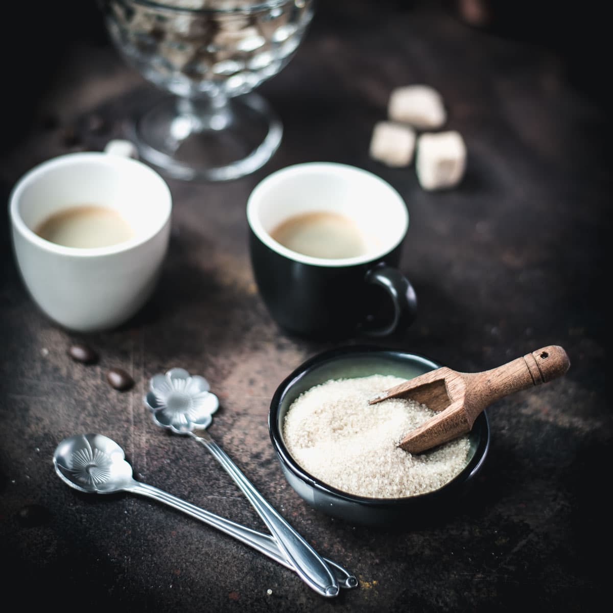 Two coffee mugs, a cup filled with brown sugar, and a glass filled with brown sugar cubes. All placed on dark ground.