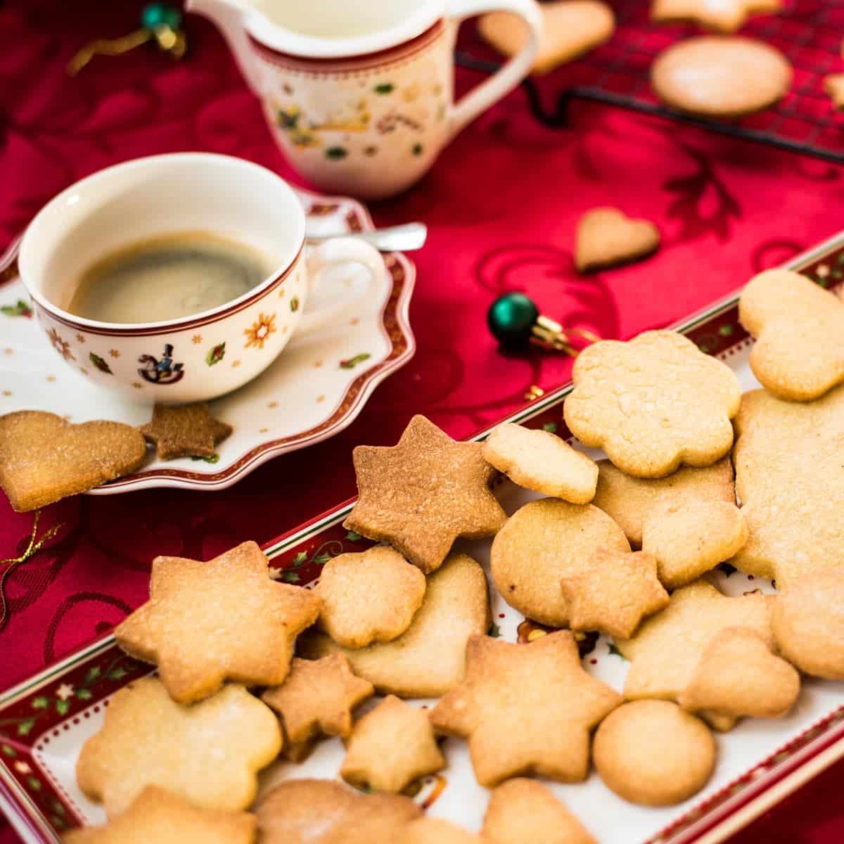 Delicious crispy sugar cookies on beautiful Christmas tray, placed on Christmassy red tablecloth.