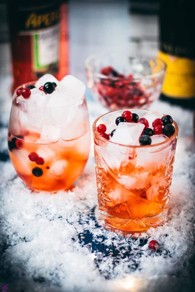 Two glasses filled with delicious cranberry Aaperol spritz, placed on snowy ground, garnished with a delicious berry mix.