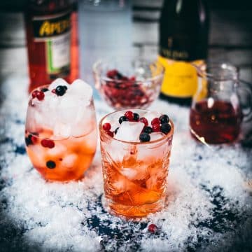 Two glasses filled with delicious cranberry Aaperol spritz, placed on snowy ground, garnished with a delicious berry mix.