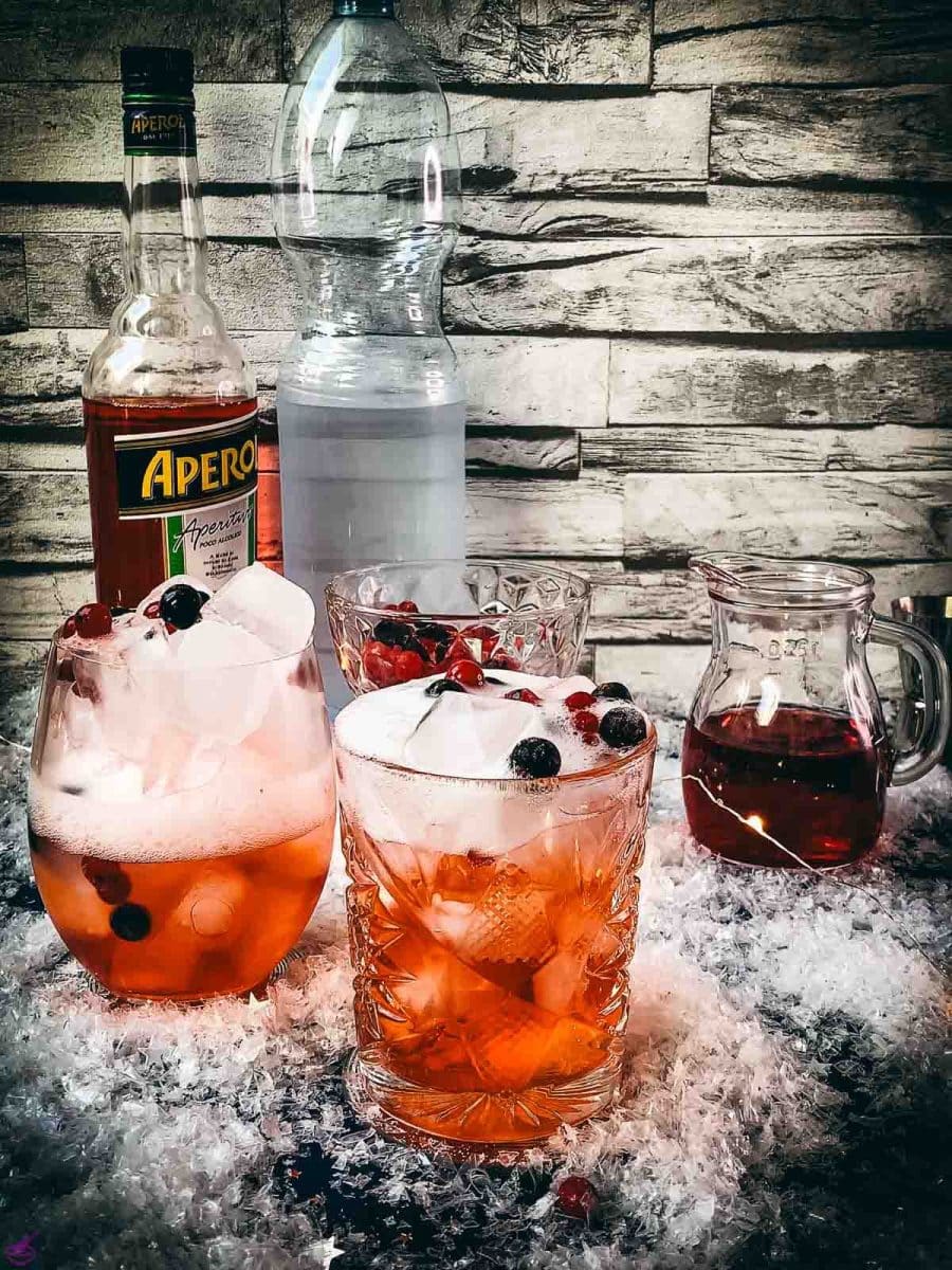 Two glasses filled with delicious cranberry Aaperol spritz, placed on snowy ground, garnished with a delicious berry mix.