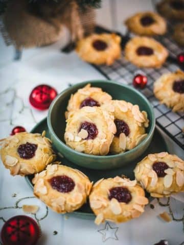 Delectable raspberry almond thumbprint cookies placed in a green bowl, next to red Christmas baubles.