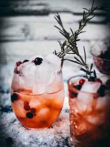 Two glasses filled with delicious cranberry Aaperol spritz, placed on snowy ground, garnished with a delicious berry mix.