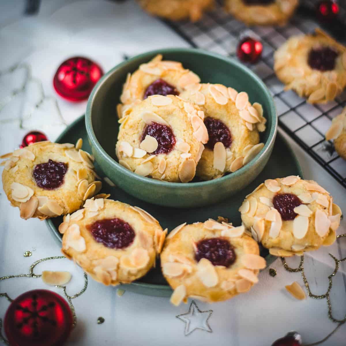 Delectable raspberry almond thumbprint cookies placed in a green bowl, next to red Christmas baubles.