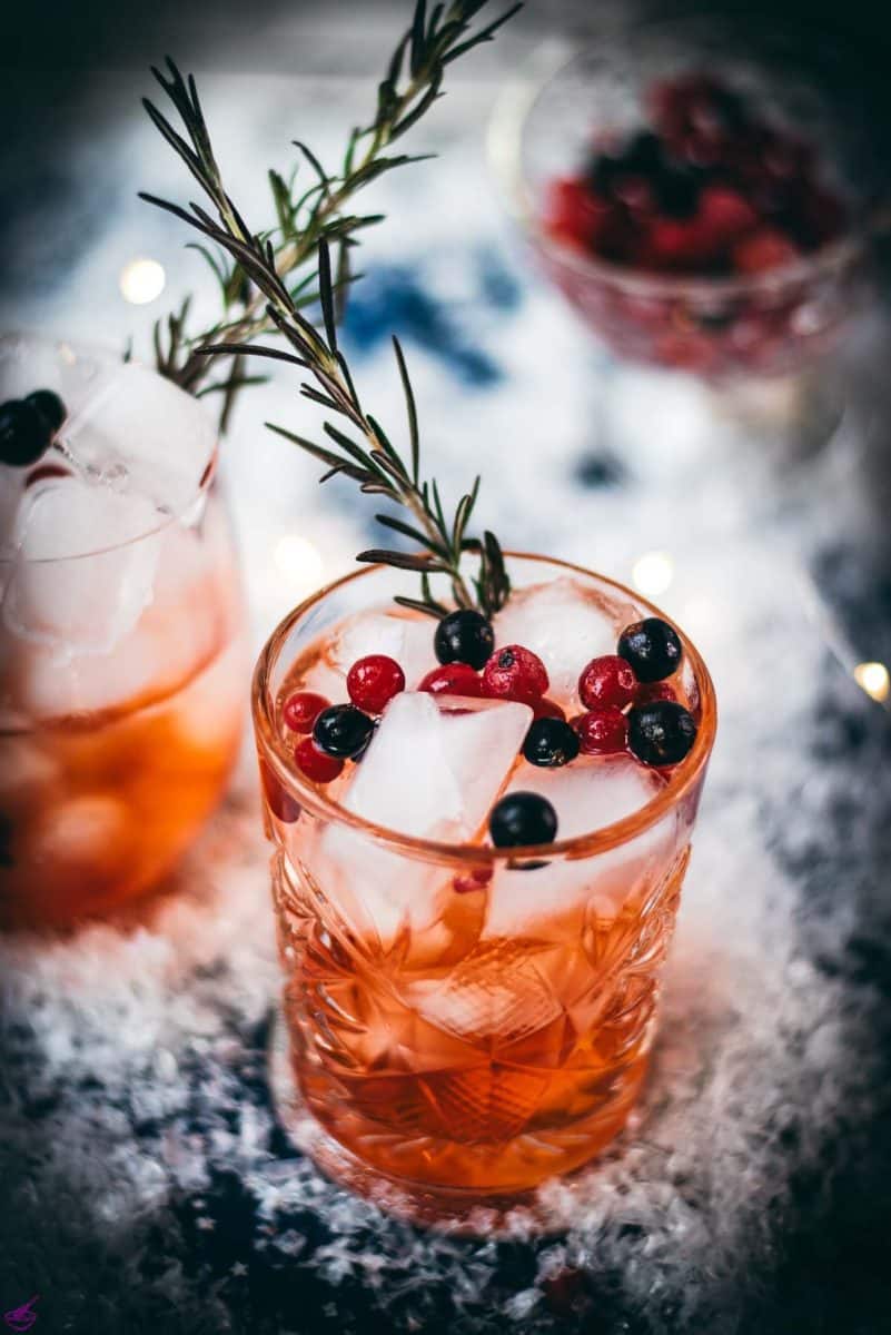 Two glasses filled with delicious cranberry Aaperol spritz, placed on snowy ground, garnished with a delicious berry mix.