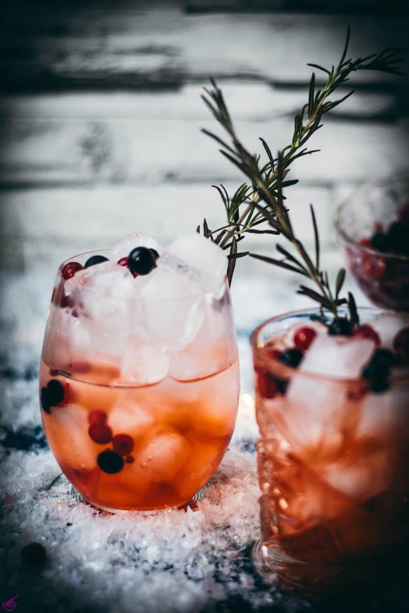 Two glasses filled with delicious cranberry Aaperol spritz, placed on snowy ground, garnished with a delicious berry mix.