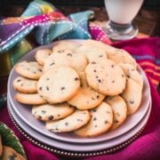 Chocolate chip sugar cookies placed on pink plates, embedded in colorful tea towels, in front of a glass of milk.