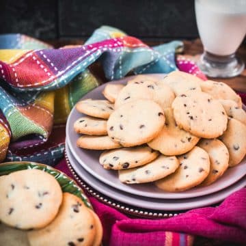 Chocolate chip sugar cookies placed on pink plates, embedded in colorful tea towels, in front of a glass of milk.