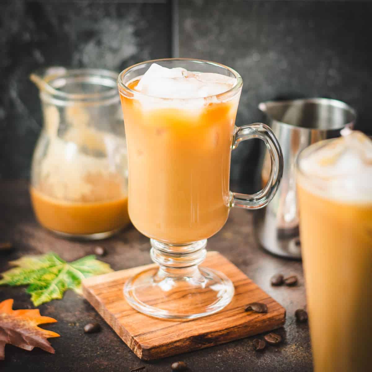 Two glasses filled with delicious iced caramel latte, placed on wooden coaster and surrounded by maple leaves.